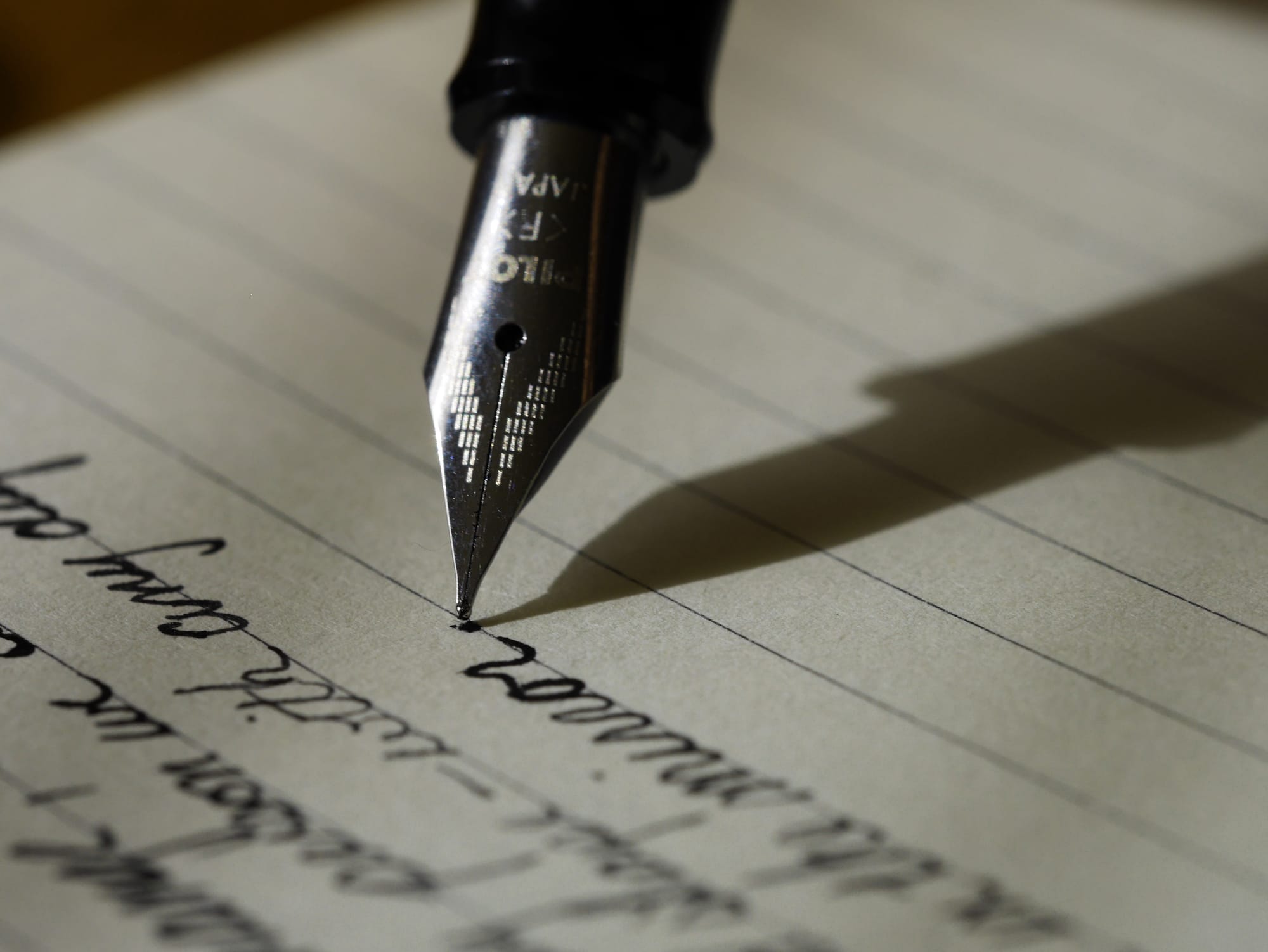 Close-up of a fountain pen nib writing on lined paper, with a soft shadow across the page.