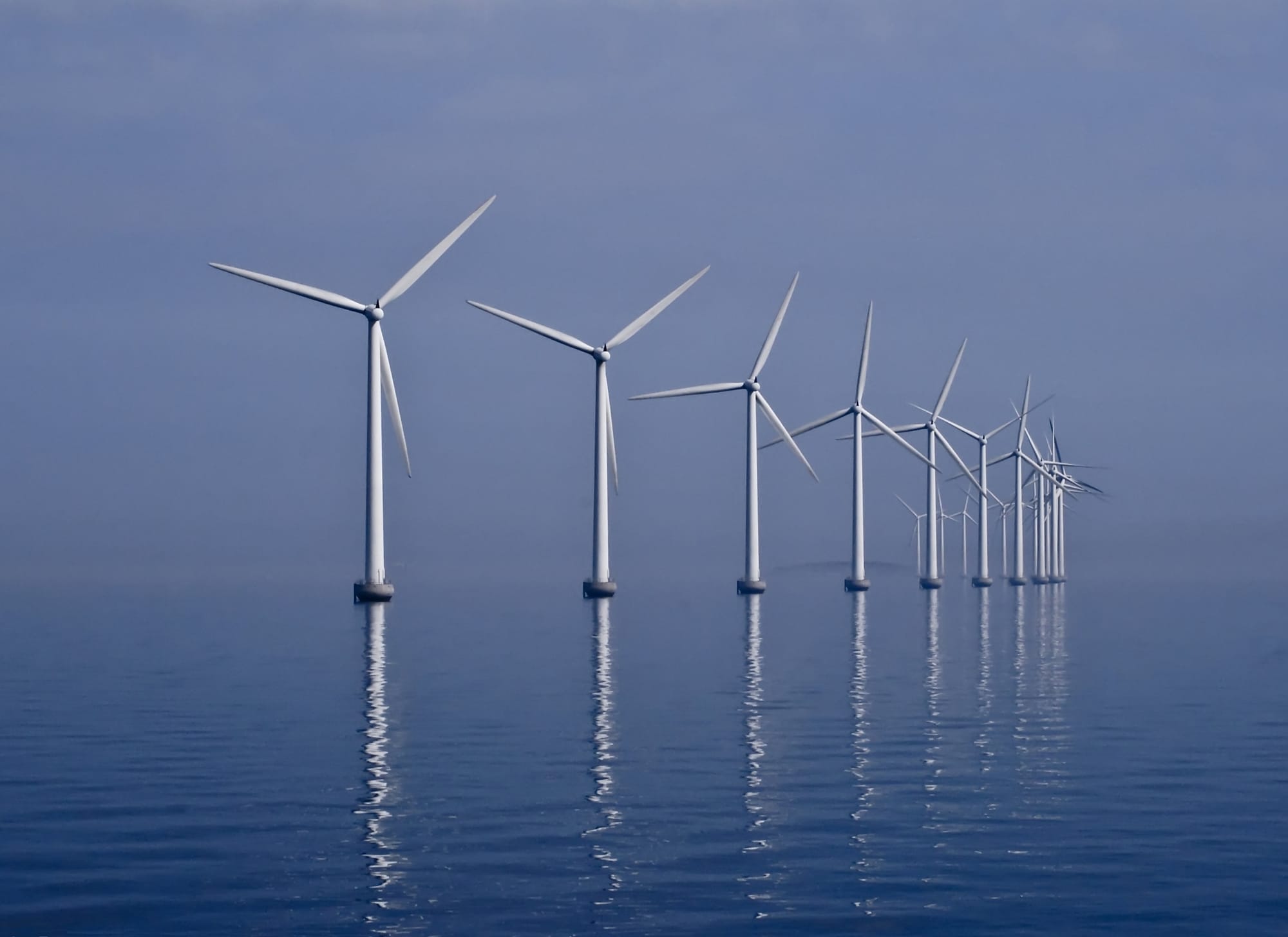 Row of offshore wind turbines at Middelgrunden near Copenhagen, reflected in calm water under a misty blue sky.
