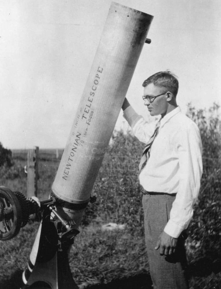 Black-and-white photo of Clyde Tombaugh standing next to his homemade 9-inch reflecting telescope in a field, circa 1930.