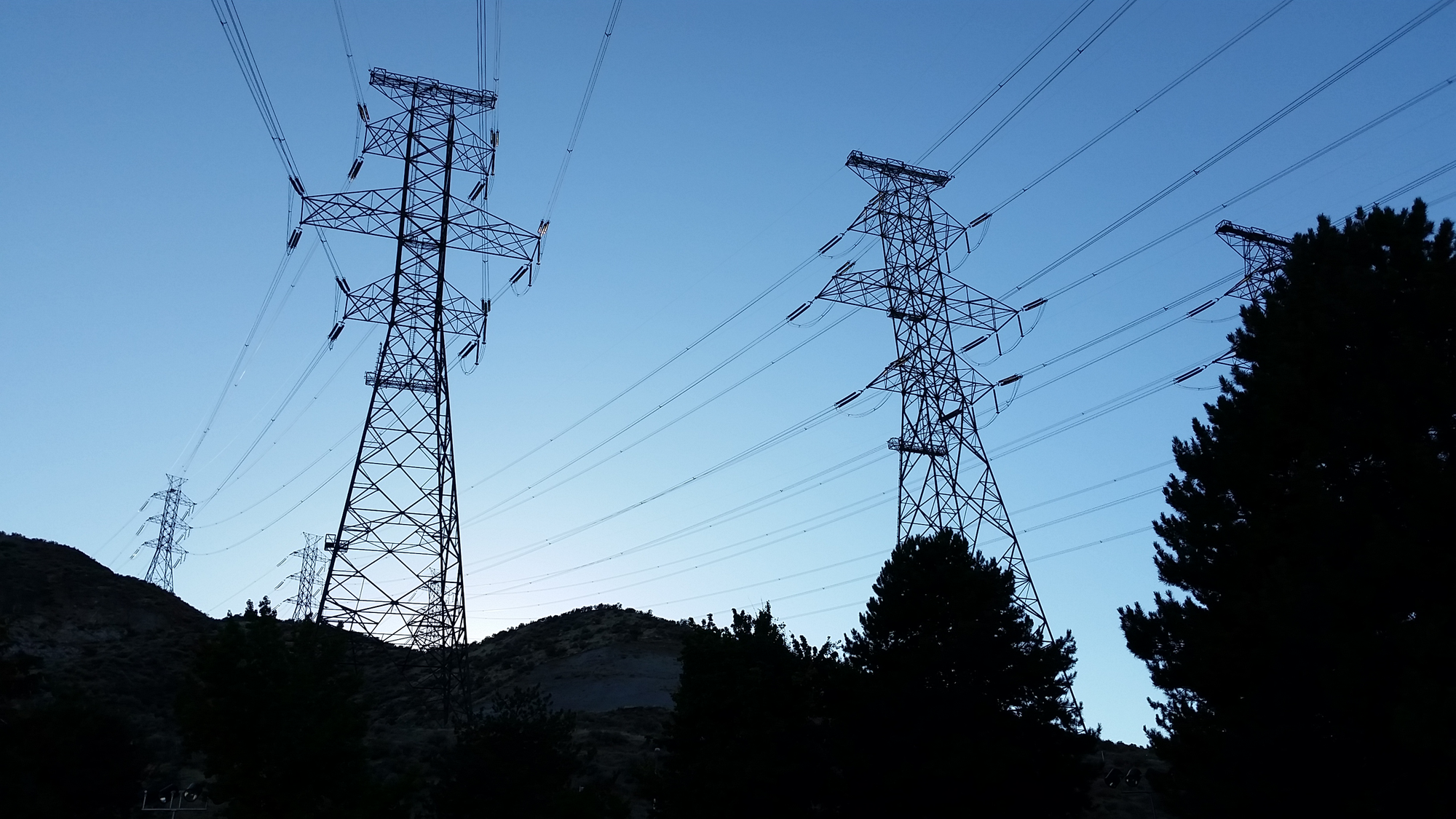 Silhouette of two large 500kV high-voltage transmission towers against a twilight blue sky, with conifer trees in the foreground.