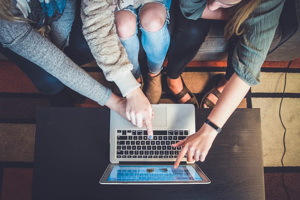 Top-down view of three people gathered around a laptop and tablet on a coffee table, fingers pointing at the screens.