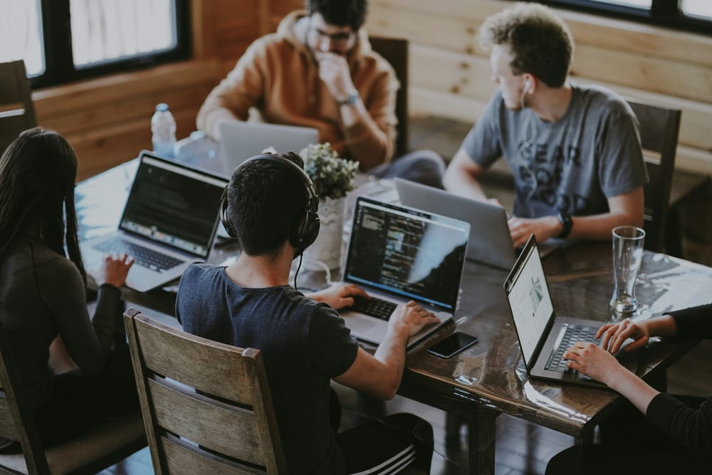 Team of developers working together on laptops around a wooden table in a sunlit workspace.