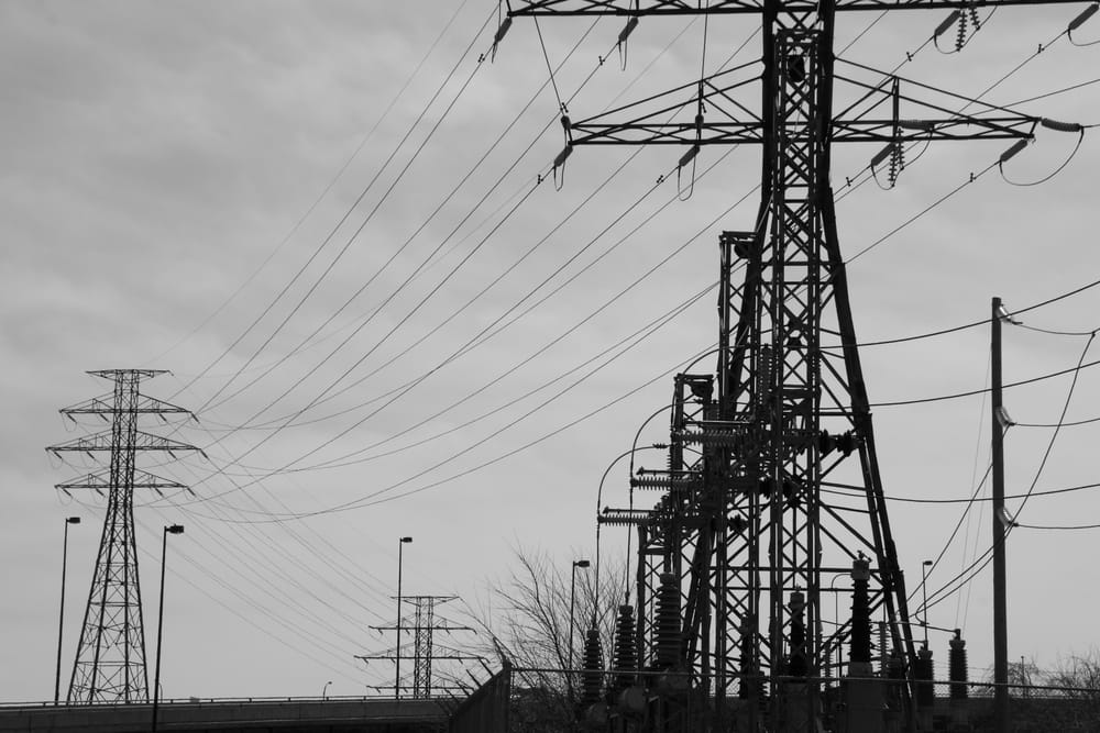 Zwart-witfoto van hoogspanningsmasten en transformatorinfrastructuur tegen een grijze lucht.