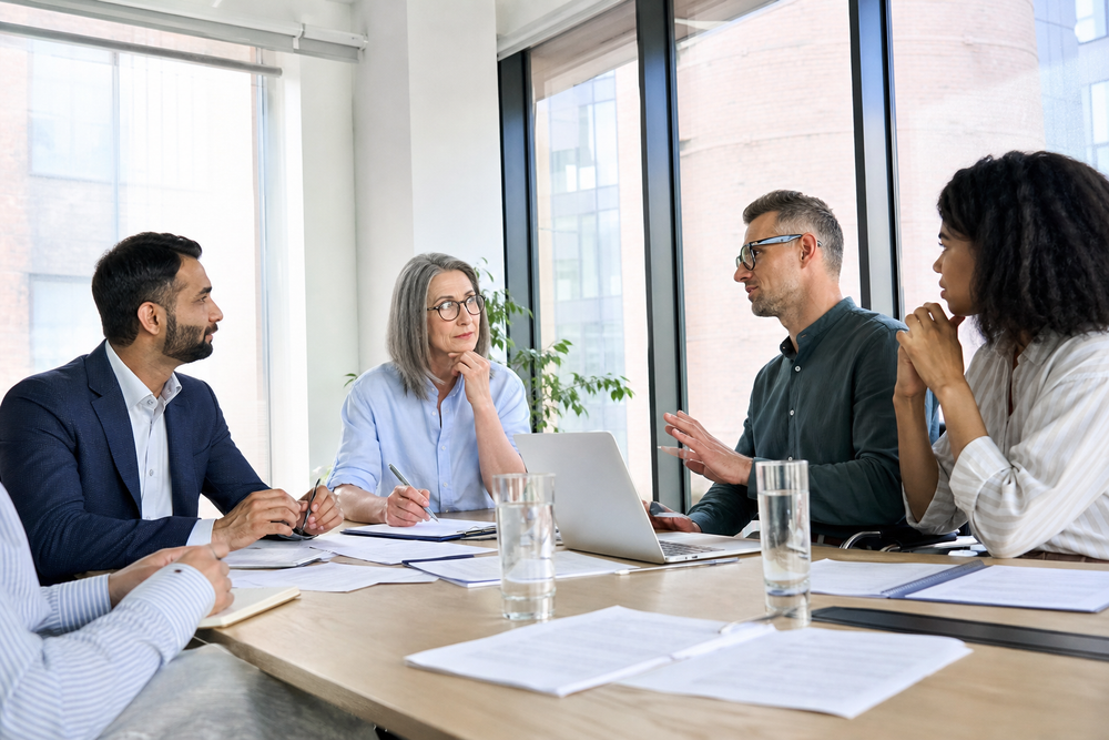 Four professionals sit around a meeting table in a bright office, discussing papers and a laptop during a serious policy conversation.