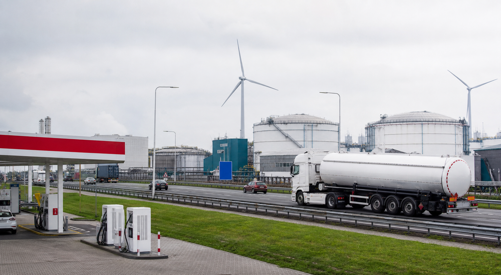 Tankstation en snelwegverkeer naast opslagtanks en windturbines onder een bewolkte lucht.